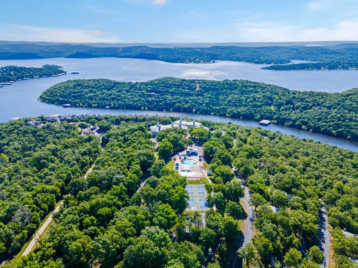 This elevated view captures the entire resort setting, featuring the shimmering lake, a sparkling swimming pool, tennis courts, and the property's buildings nestled among lush greenery. The image radiates a sense of tranquility while showcasing the variety of activities available, from boating and swimming to tennis and hiking.