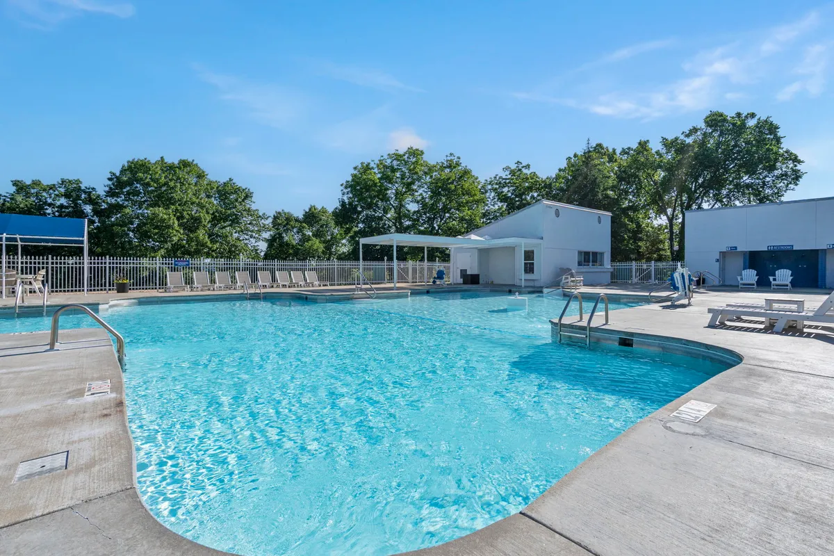 A vibrant outdoor scene showing a sunlit swimming pool surrounded by comfortable loungers and a poolside cabana. The atmosphere is leisurely, with groups and individuals enjoying the water, sunbathing, and socializing by the pool.