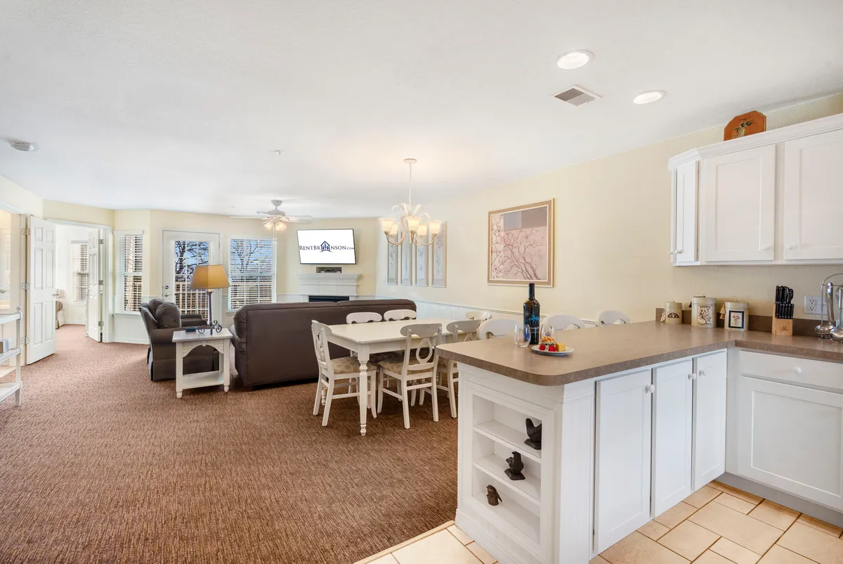 This image showcases a spacious, open-concept living area that seamlessly connects to the kitchen and dining spaces. A kitchen island, dining table, and lounge area with a television are all visible, emphasizing the easy flow between these zones. The warm lighting and comfortable furnishings create a welcoming atmosphere ideal for socializing or relaxing as a group.