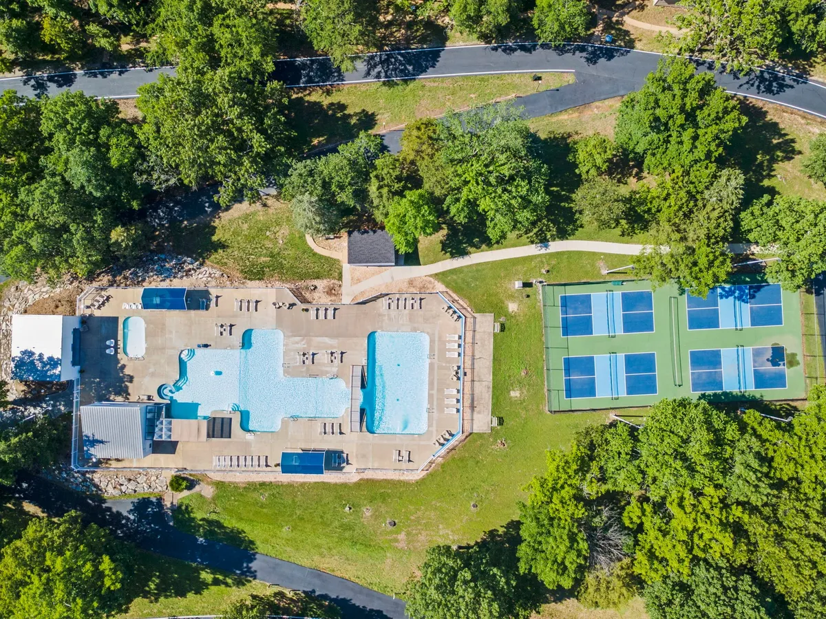 A vibrant outdoor scene featuring a sparkling swimming pool surrounded by lounge chairs, tennis courts in the background, and leafy trees along the perimeter. The pool area is sunlit, inviting relaxation and play, while the visible courts hint at additional recreational options.