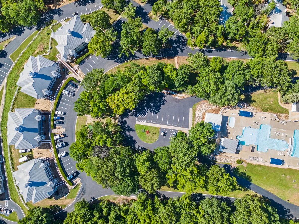An exterior shot of the swimming pool surrounded by trees, with the building and parking area in view. The scene is bright and welcoming, promising relaxation and fun in the sun during summer months.