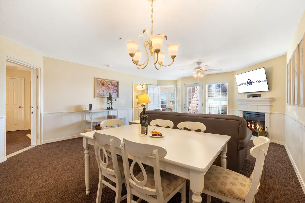 A cozy dining table sits beneath a chandelier, with a warm fireplace and television nearby. The space is arranged for gatherings, with the dining and living areas closely integrated, creating a perfect setting for family meals and relaxed evenings by the fire.