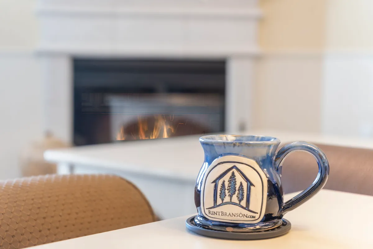 A close-up of a ceramic mug on a table with a glowing fireplace in the background, evoking a sense of warmth and relaxation. The scene captures the mood of enjoying quiet, comfortable moments at home, whether alone or with loved ones.
