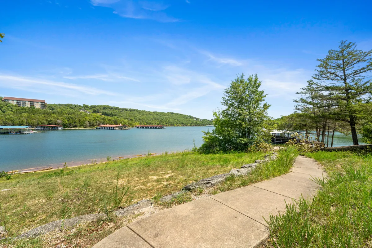 A tranquil lakeside scene featuring a tree-lined pathway winding toward the water. The lake glistens in the background, surrounded by hills, inviting morning strolls, dog walks, or spontaneous dips in the water.
